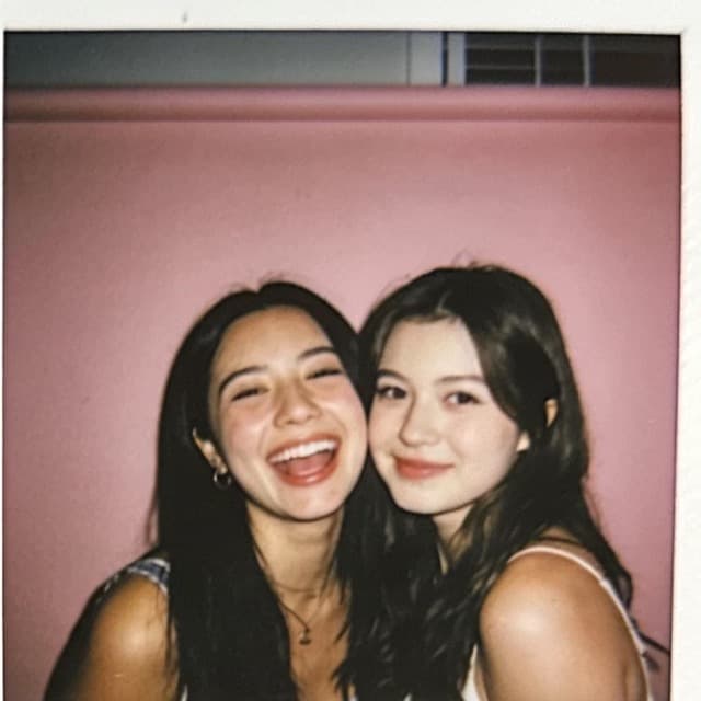 The same two friends grinning against a pink wall
